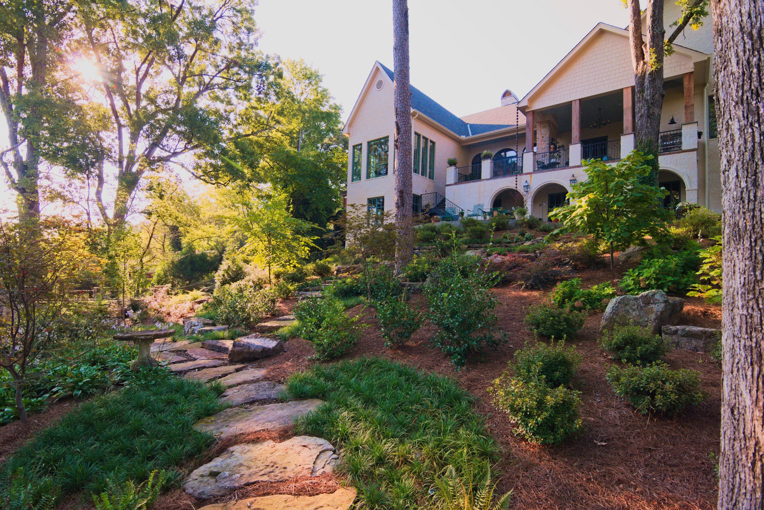 A large, elegant house with tall windows and a spacious balcony overlooks a landscaped garden with stone steps, lush greenery, and mature trees in the warm sunlight.
