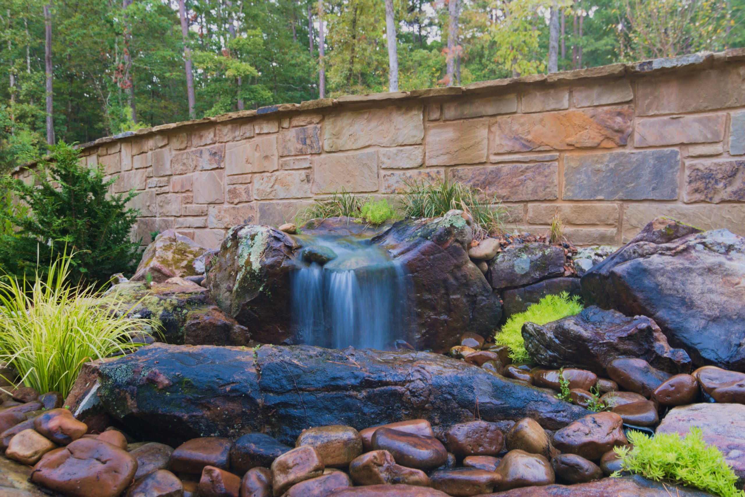 A small waterfall flows over mossy rocks into a bed of smooth stones, surrounded by green plants, with a stone wall and trees in the background.