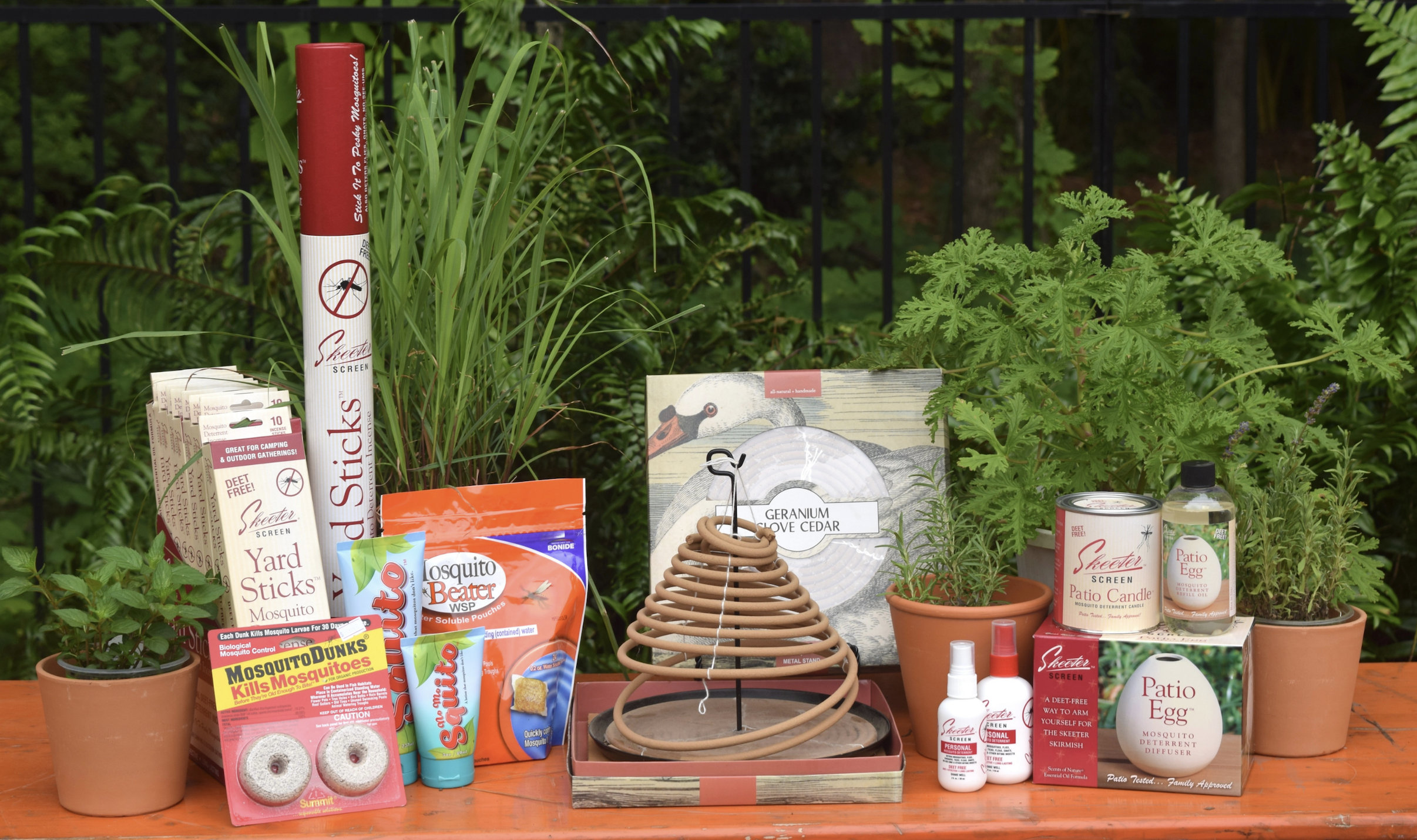 A collection of mosquito control products and potted plants displayed on an orange table, including candles, yard sticks, coils, sprays, and granules, set outdoors with green foliage in the background.