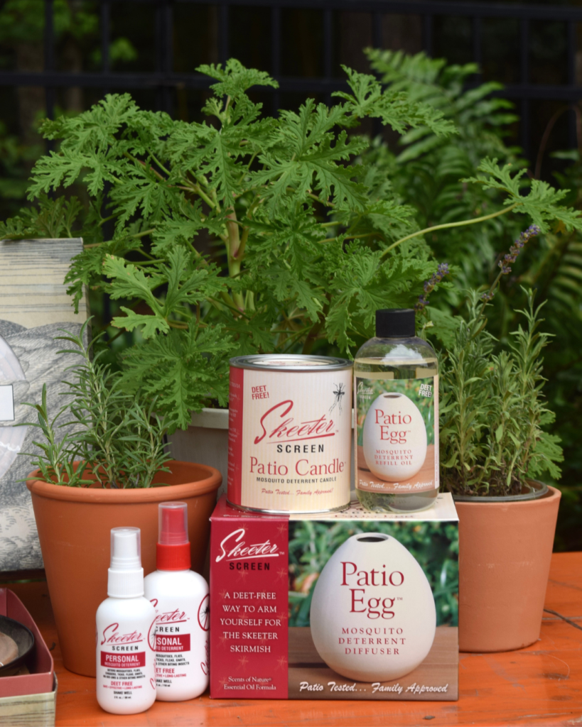 A collection of mosquito repellent products, including sprays, a patio candle, and a patio egg diffuser, are displayed in front of potted plants on an outdoor surface.