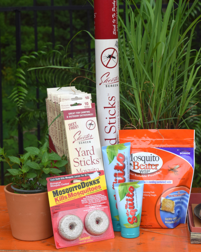 A collection of mosquito control products, including sprays, granules, repellent sticks, mosquito dunks, and yard sticks, is arranged on an orange table with green plants in the background.
