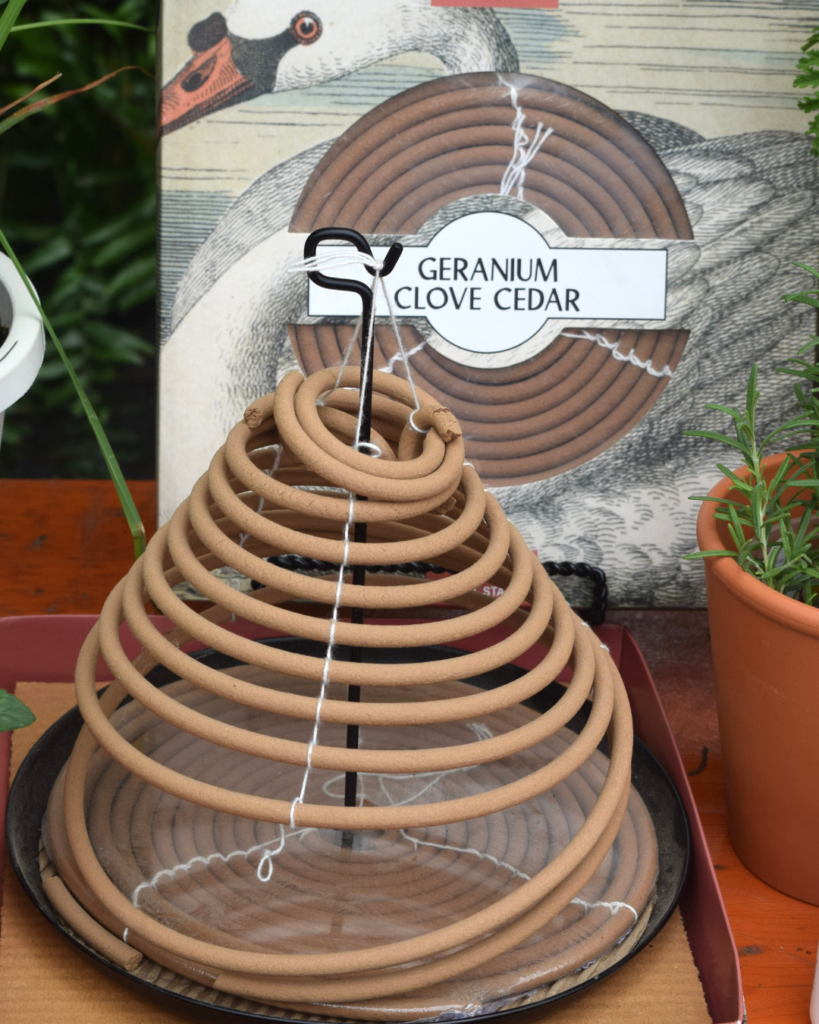 A large spiral mosquito coil labeled Geranium Clove Cedar sits on a metal tray, with a potted plant nearby and an illustrated bird in the background.
