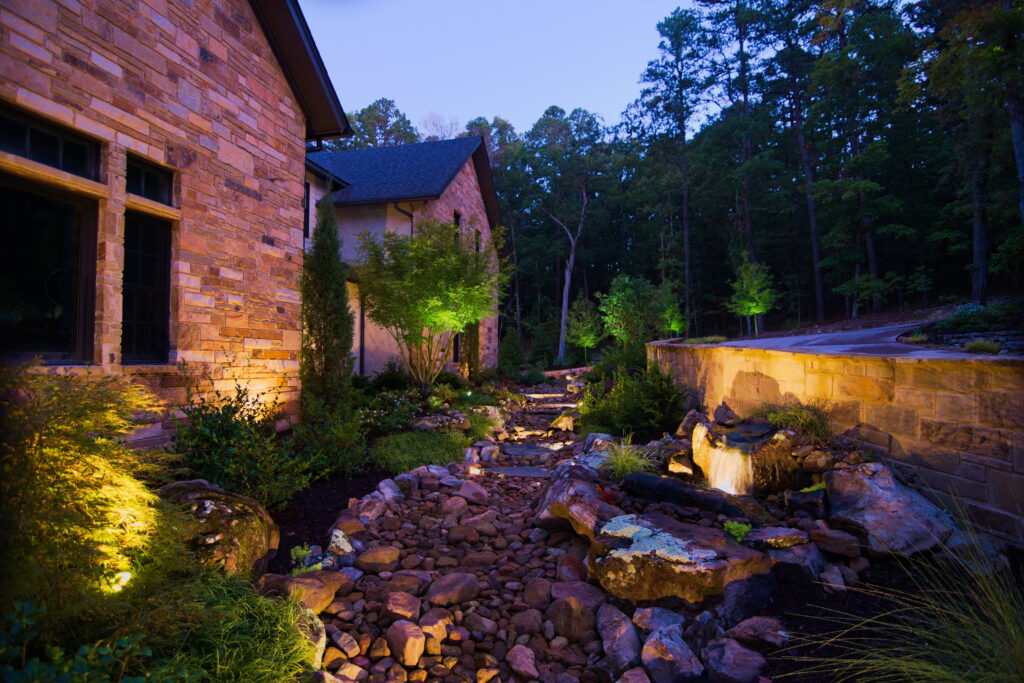 A landscaped backyard at dusk features a stone path, waterfall, and lush greenery with landscape lighting softly illuminating the garden beside a brick house, bordered by a stone wall and surrounded by tall trees.