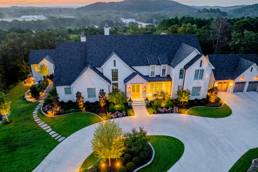 Aerial view of a large, elegant white house with multiple gables, manicured lawns, and gardens, beautifully enhanced by landscape lighting at dusk, with wooded hills in the background.