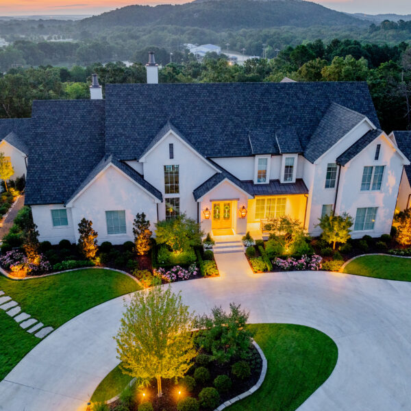 Aerial view of a large, elegant white house with multiple gables, manicured lawns, and gardens, beautifully enhanced by landscape lighting at dusk, with wooded hills in the background.