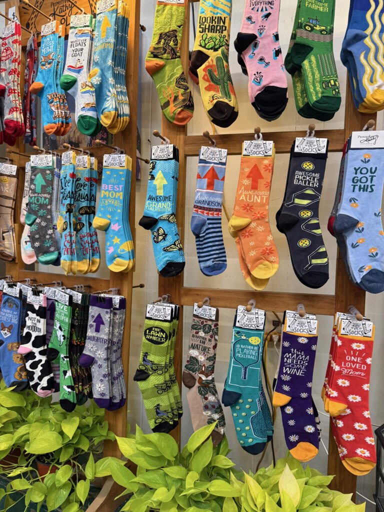 A display of colorful socks with fun patterns and humorous phrases is arranged on a wooden rack above green leafy plants—perfect gifts ideas for gardeners. Sock designs feature animals, stars, and witty text for various occasions.