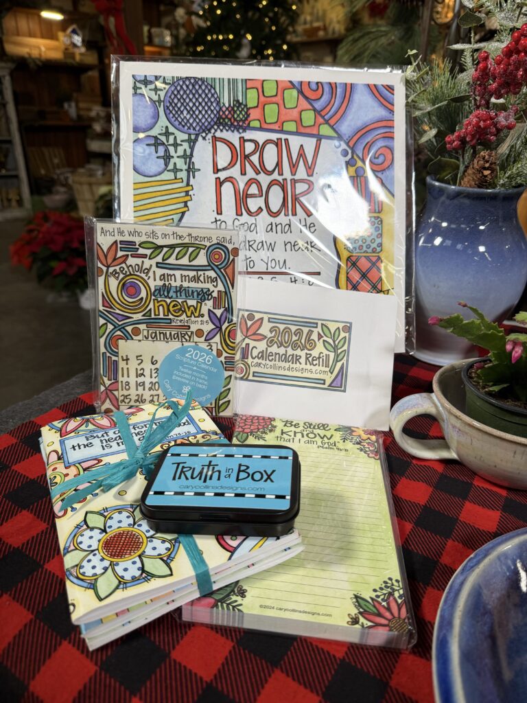 A colorful 2026 calendar refill set with hand-drawn designs, an inspirational card, a Truth Box tin, and a notepad from an Arkansas gift shop, all displayed on a red and black plaid tablecloth with plants and a teacup nearby.