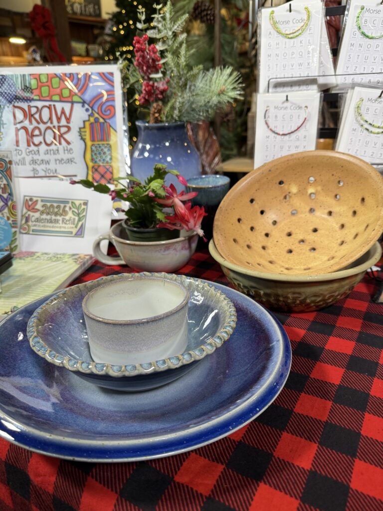 Handmade pottery bowls, plates, and a cup are displayed on a red and black plaid tablecloth at an Arkansas gift shop, with a decorative plant, calendar refills, and a “Draw Near” sign in the background.