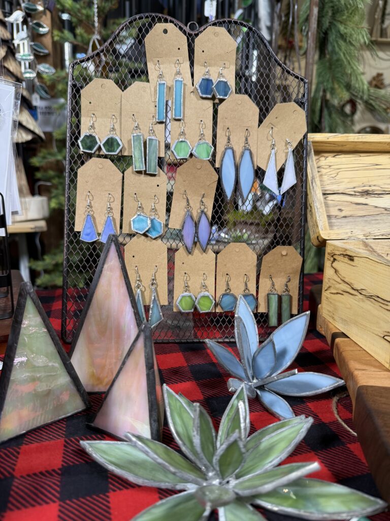 A display of stained glass earrings in blue and green tones hangs on a mesh stand, surrounded by geometric shapes and flowers on a red and black plaid tablecloth—perfect for any Arkansas gift shop.