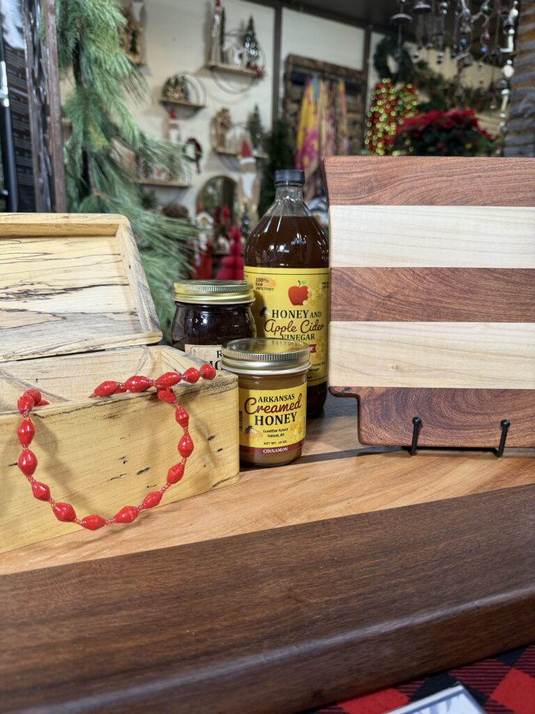 A charming Arkansas gift shop display features wooden boxes and cutting boards, a red beaded necklace, a jar of creamed honey, honey apple cider vinegar, and festive holiday decor in the background.