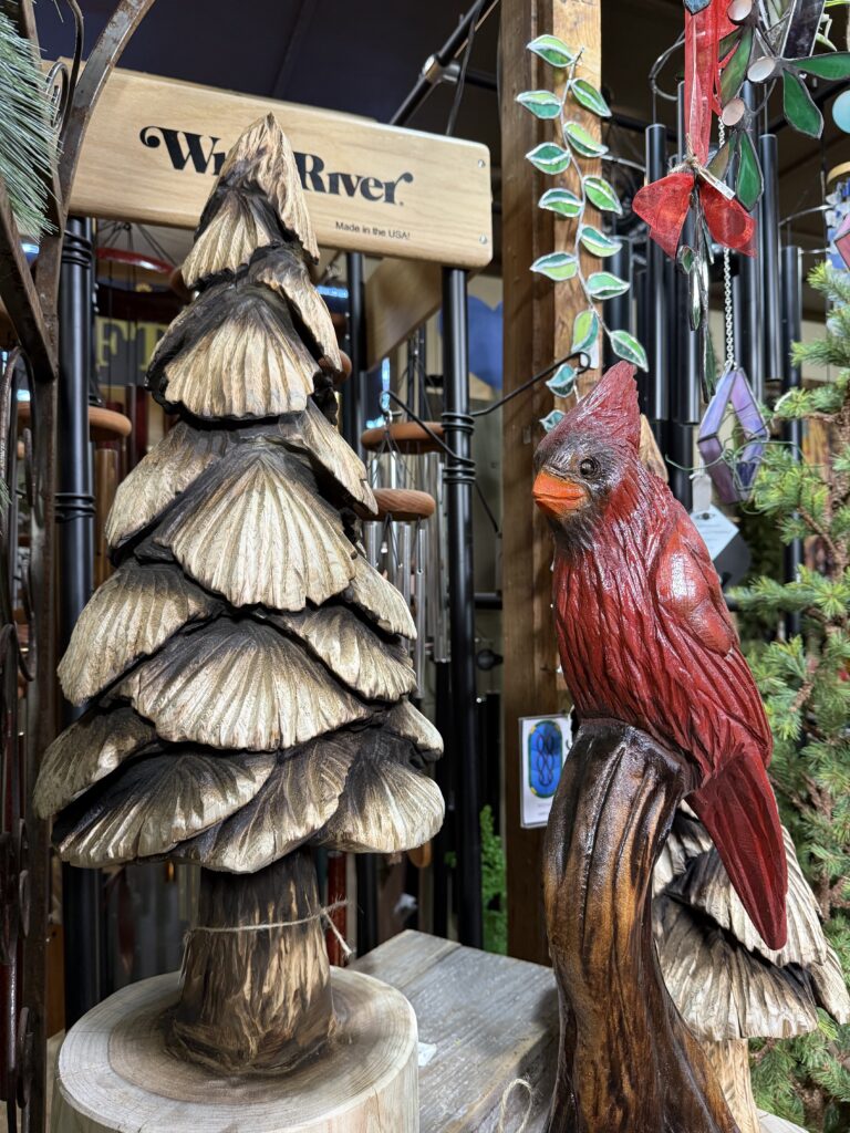 A wooden sculpture of a red cardinal perched on a branch stands next to a carved tree, surrounded by colorful garden ornaments near an Arkansas gift shop and a sign that reads White River Made in the USA in the background.