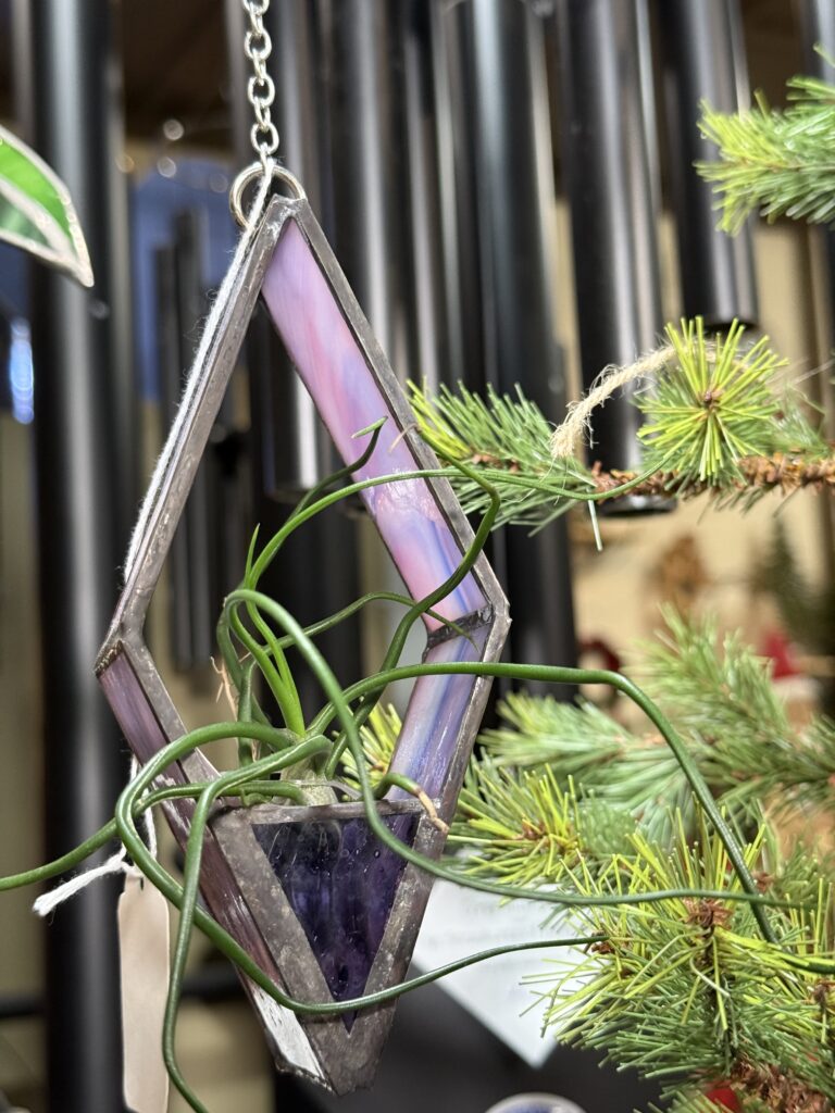 A geometric, diamond-shaped stained glass hanging planter from an Arkansas gift shop features purple and clear sections and holds a small green air plant. It is displayed on an artificial pine branch with wind chimes blurred in the background.