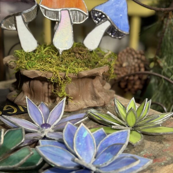 Stained glass sculptures of mushrooms and succulents are displayed on a wooden surface with moss and pinecones in the background, brightly lit by sunlight—an enchanting find at an Arkansas gift shop.