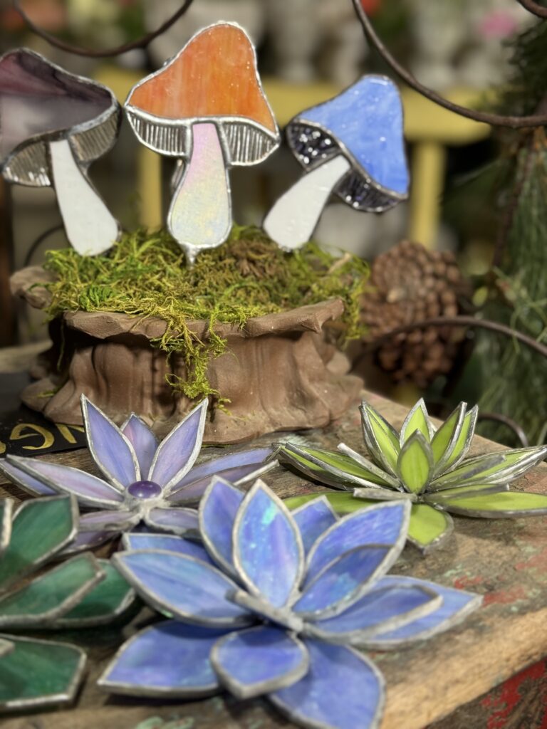 Stained glass sculptures of mushrooms and succulents are displayed on a wooden surface with moss and pinecones in the background, brightly lit by sunlight—an enchanting find at an Arkansas gift shop.