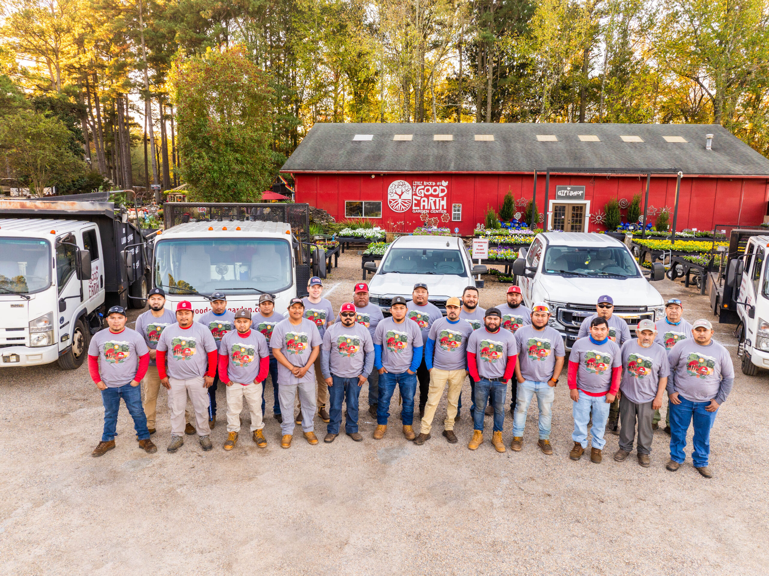 A group of workers in matching gray shirts stand in front of several trucks and a red building labeled Good Earth. The area is surrounded by trees and plants, and the group is smiling at the camera.