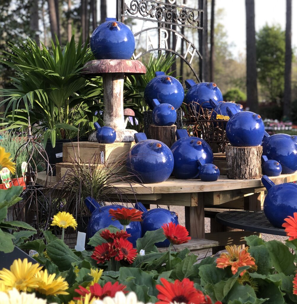 A garden display features numerous shiny blue ceramic spheres on wooden tables, surrounded by colorful blooming flowers and green plants, with a decorative metal sign visible in the background.