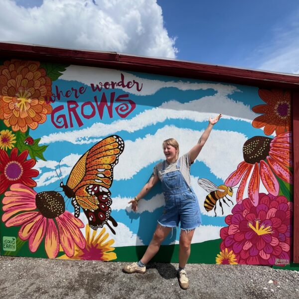 A smiling person in denim overalls stands with arms outstretched in front of a colorful mural featuring flowers, butterflies, and the words where wonder GROWS painted on a wall under a partly cloudy sky.