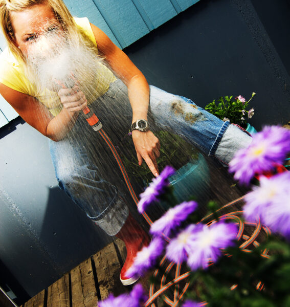 A woman in jeans and a yellow shirt crouches on a wooden deck, spraying water from a hose toward purple flowers, with water splashing in front of her face.