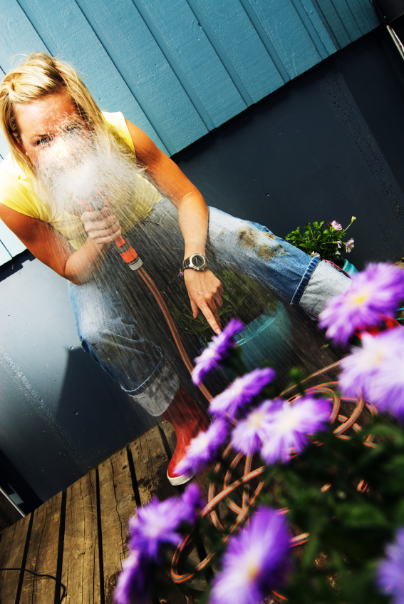 A woman in jeans and a yellow shirt crouches on a wooden deck, spraying water from a hose toward purple flowers, with water splashing in front of her face.