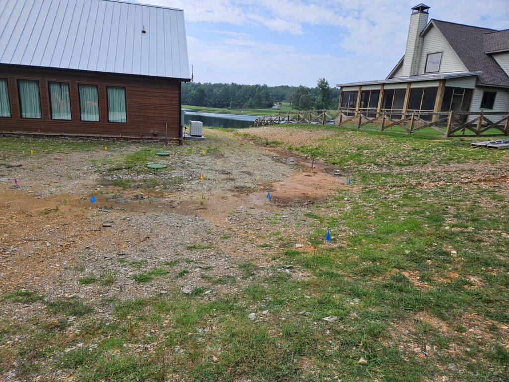 A grassy and gravel area with colored utility flags between two houses near a lake hints at future garden transformations; one house has a metal roof, the other a chimney and porch with a wooden fence, all set against trees and water under a partly cloudy sky.