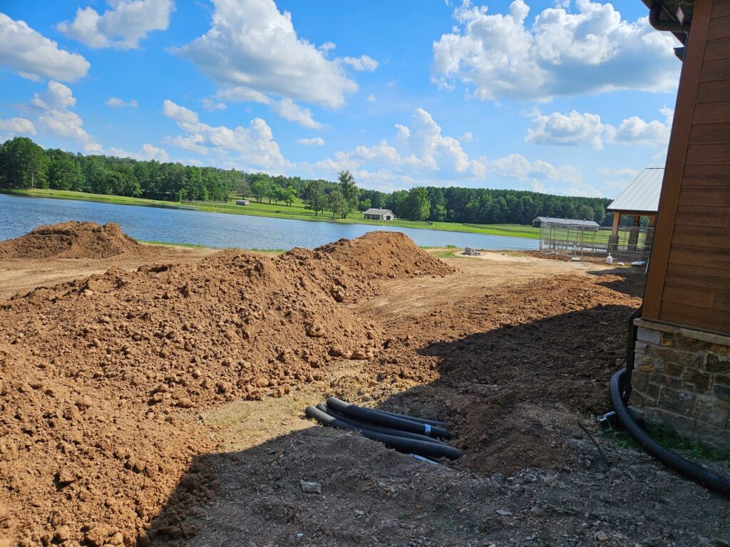 A construction site with large dirt piles and black pipes lies next to a scenic lake, surrounded by trees under a blue sky. On the right, a building with a stone foundation hints at future garden transformations in this tranquil setting.