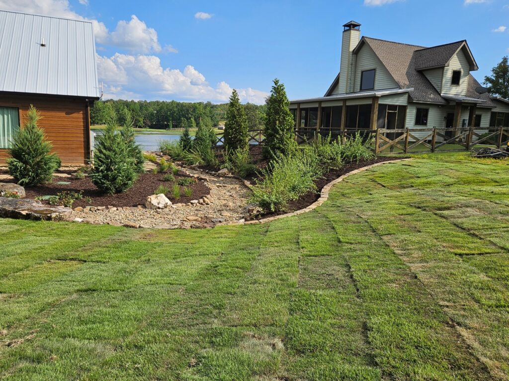 A house with a metal roof and a screened porch sits beside a landscaped yard, showcasing garden transformations with newly laid sod, small trees, shrubs, mulch, and rocks—all overlooking a lake and forest under a blue sky.
