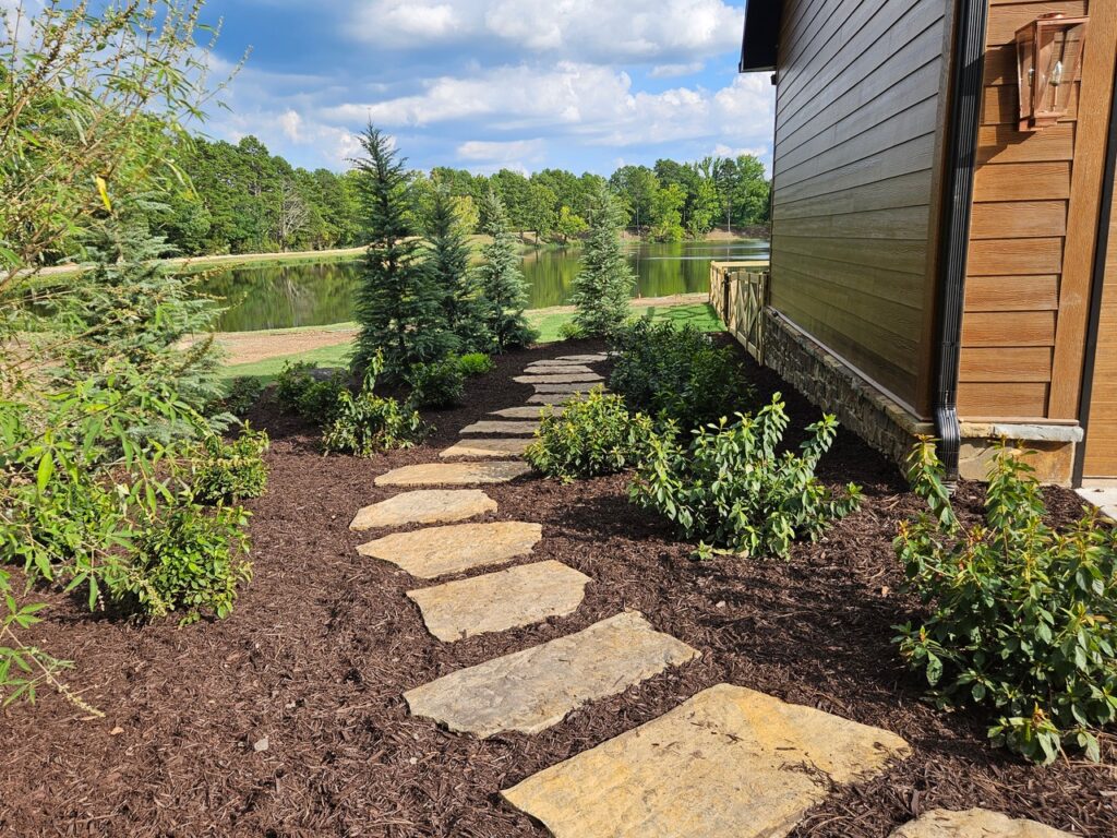 Stone path with irregular stepping stones leads through landscaped garden beds, showcasing garden transformations with mulch, shrubs, and young trees beside a wooden building, overlooking a pond and green trees under a partly cloudy sky.