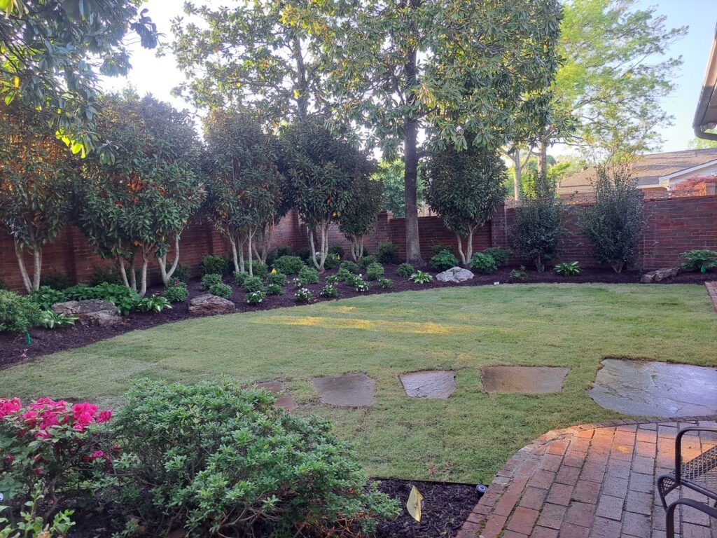 Experience stunning garden transformations in this neatly landscaped backyard, where a grassy lawn, stone pavers, and vibrant flowerbeds border a red brick wall beneath tall trees. Sunlight filters through, casting gentle shadows over the serene garden.