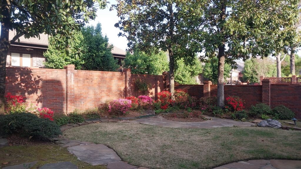 A backyard showcasing stunning garden transformations with a curved stone walkway, grassy area, blooming pink and red flowers along a red brick wall, leafy green bushes, and tall trees providing shade.