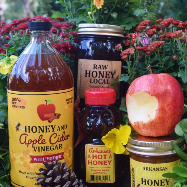 A display of honey products, apple cider vinegar, an apple, and a pine cone arranged among yellow and red flowers outdoors. Labels read “Honey and Apple Cider Vinegar”, “Raw Honey”, “Arkansas Hot Honey”, and “Arkansas Honey Lemon.”.