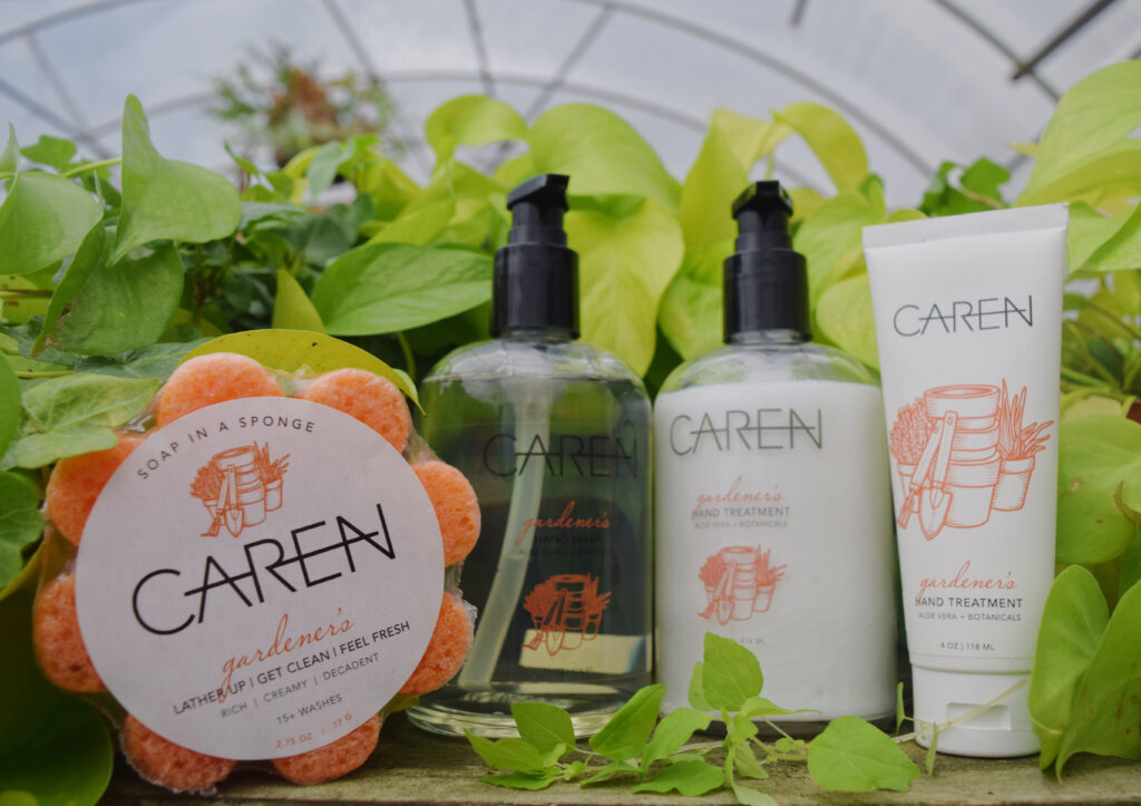 A set of CARĒN branded skincare products, including soap in a sponge, two pump bottles, and a tube, arranged on a wooden surface with green leafy plants in the background.