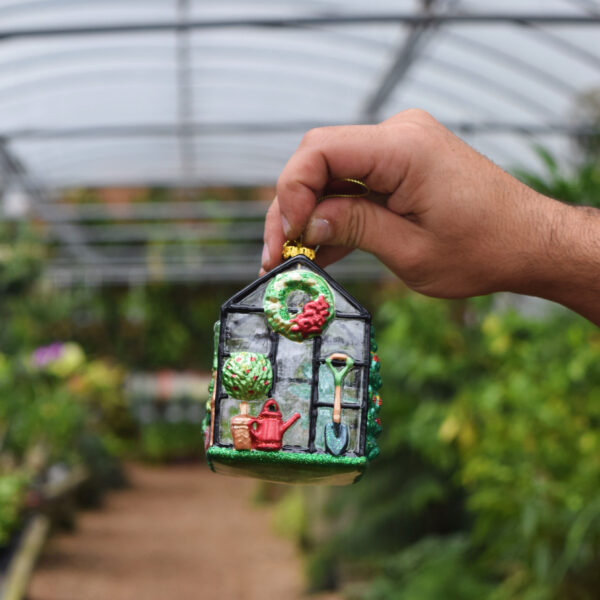 A hand holds a small, colorful greenhouse ornament decorated with gardening tools and plants, with a blurred background of green plants inside a greenhouse.