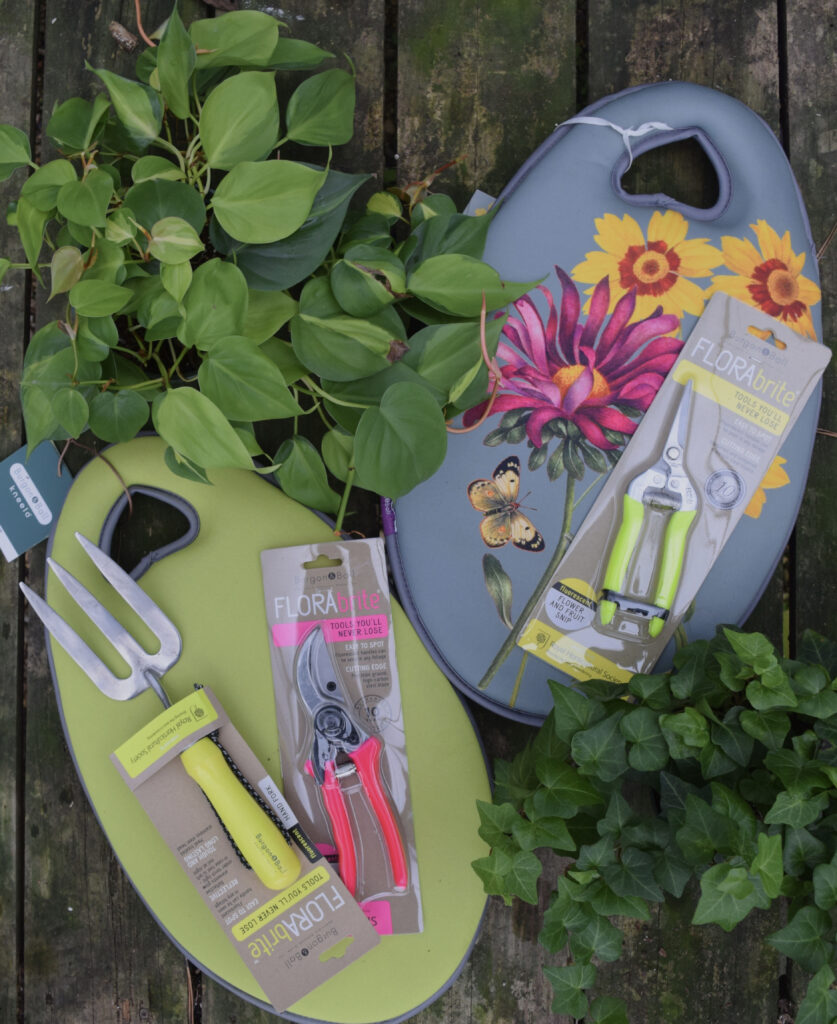 Two kneeling pads, one green and one gray with a floral butterfly design, are on a weathered wooden surface. Green garden plants, a hand fork, pruners, and a weeder are arranged around them.