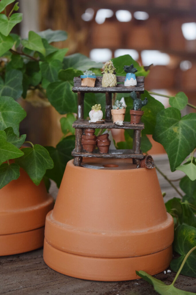 A small wooden shelf filled with miniature potted succulents sits on top of two stacked terracotta pots, surrounded by green ivy leaves.