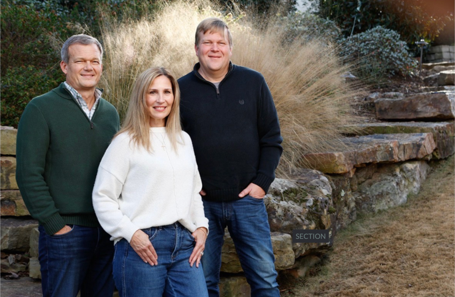 Three adults, two men and one woman, stand outdoors in front of stone steps and tall grass. They are smiling and wearing casual sweaters and jeans. The setting appears to be a landscaped garden or park.