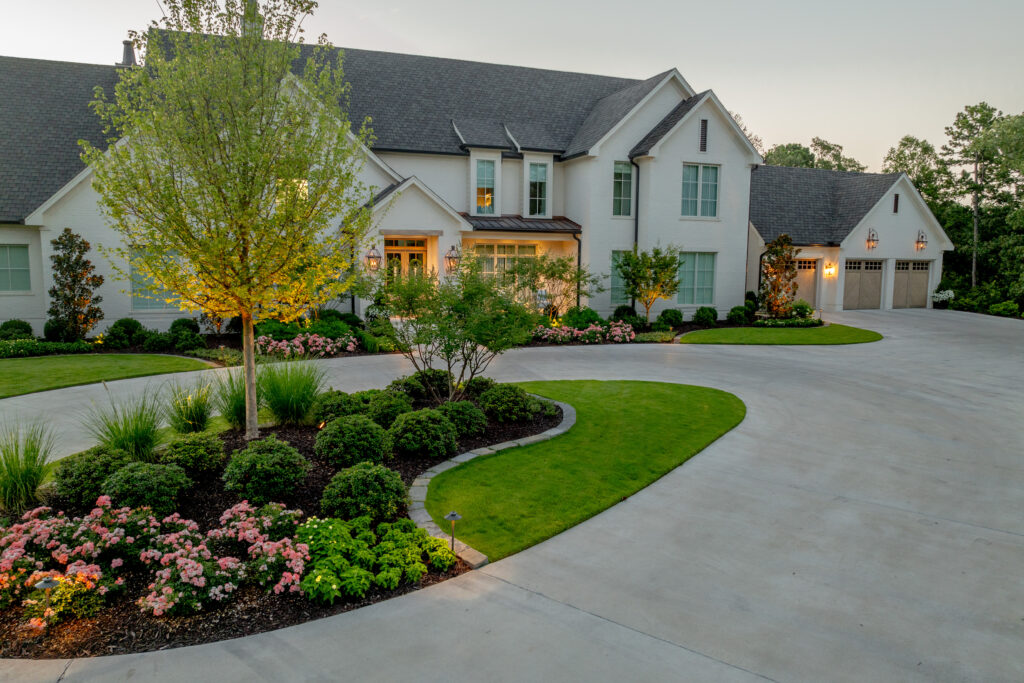 A large, elegant house with white exterior walls, tall windows, and a well-manicured lawn. Flower beds, shrubs, and trees line the curved driveway leading to a three-car garage. The scene is lit softly at dusk, all showing the best plants for landscaping 