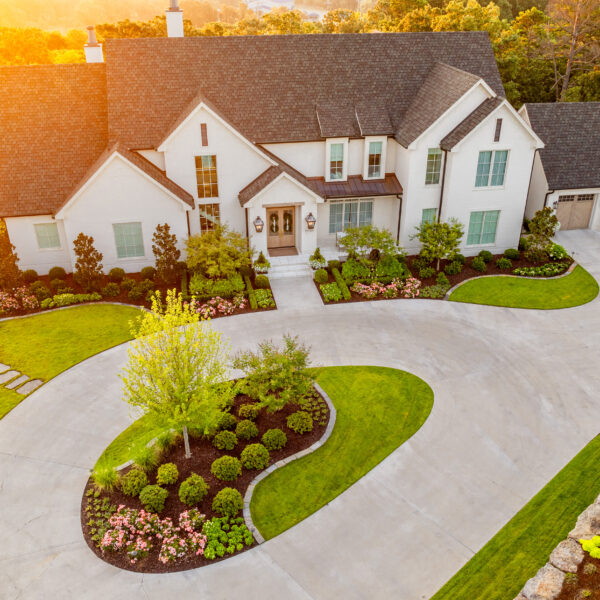 Aerial view of a large, two-story white house with gray roof, multiple gables, three-car garage, circular driveway, and manicured lawn surrounded by landscaped gardens and trees at sunset.