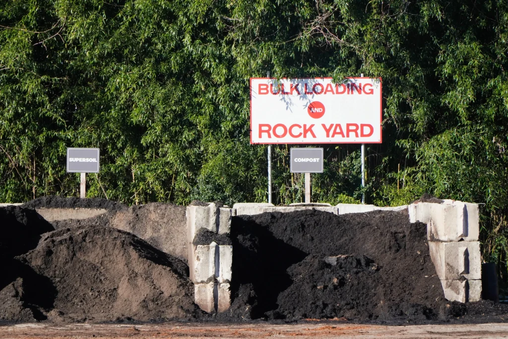 Three large piles of dark soil materials are separated by concrete barriers. Behind them are signs reading “SUPERSOIL,” “COMPOST,” and a large “BULK LOADING AND ROCK YARD” sign, with green trees in the background.