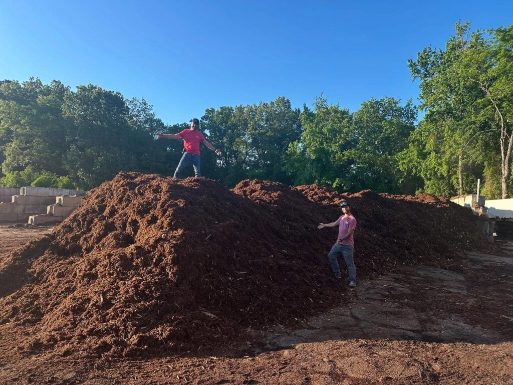 Two people stand on a large pile of landscaping materials at a garden center, surrounded by trees and plants for landscaping under a clear blue sky.