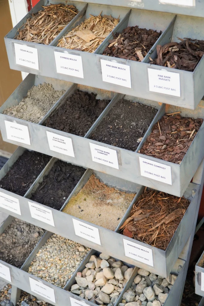 A display rack holds metal bins filled with different types of mulch, soil, sand, and gravel, each labeled clearly, showing a variety of landscaping materials for sale.