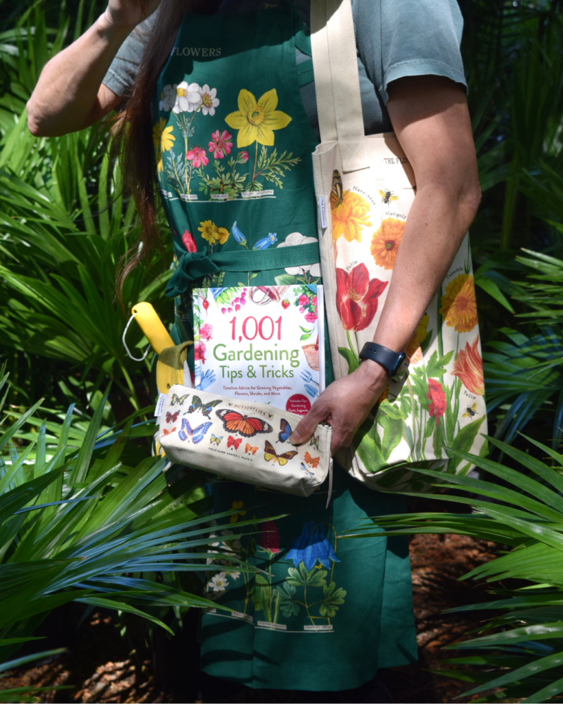 A person in a green floral apron holds a tote bag with colorful flowers, a gardening book titled 1,001 Gardening Tips & Tricks, and a butterfly-patterned pouch, standing among green plants.