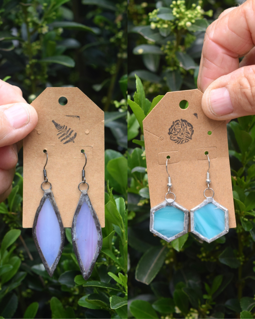 Two hands each hold a brown tag displaying stained glass earrings against green foliage. The left tag has purple leaf-shaped earrings; the right tag has blue hexagonal earrings. Both tags feature stamped designs.