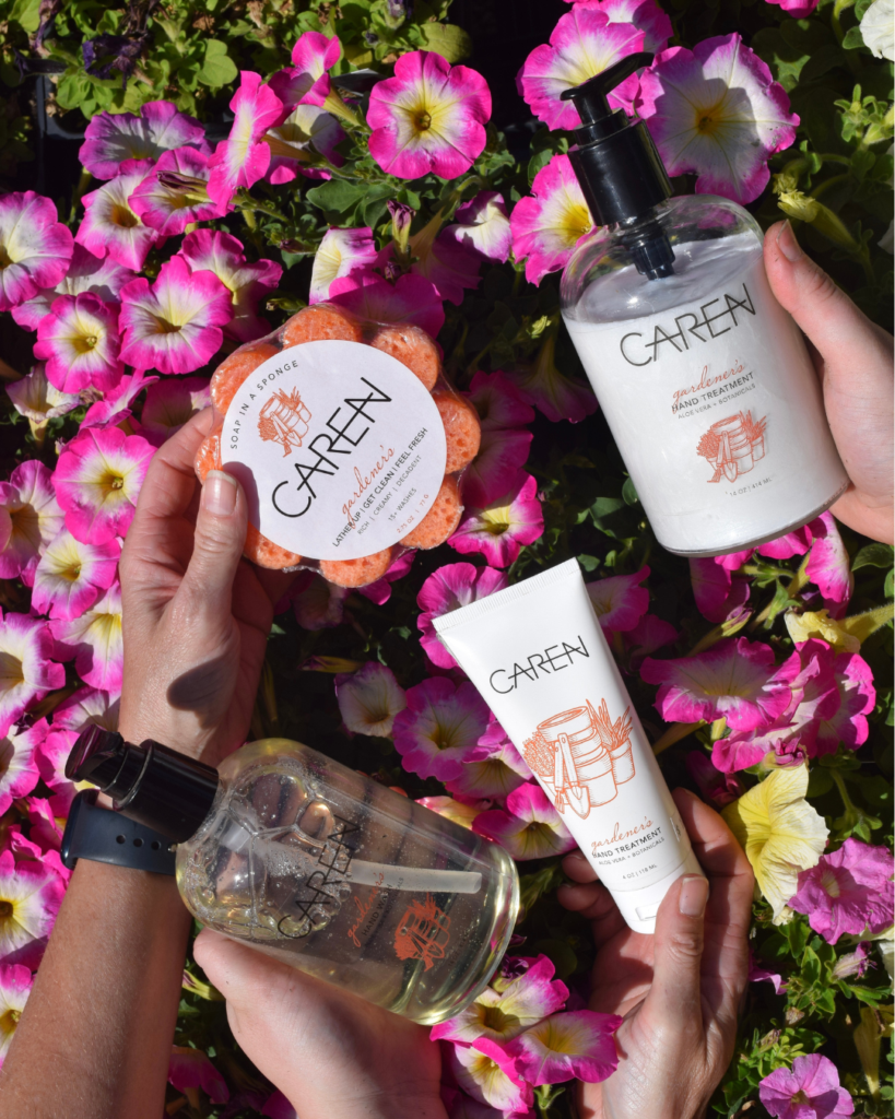 Three hands hold CAREN brand skincare products—a lotion, a body wash, a hand cream, and a sponge—against a vibrant background of blooming pink and white flowers.
