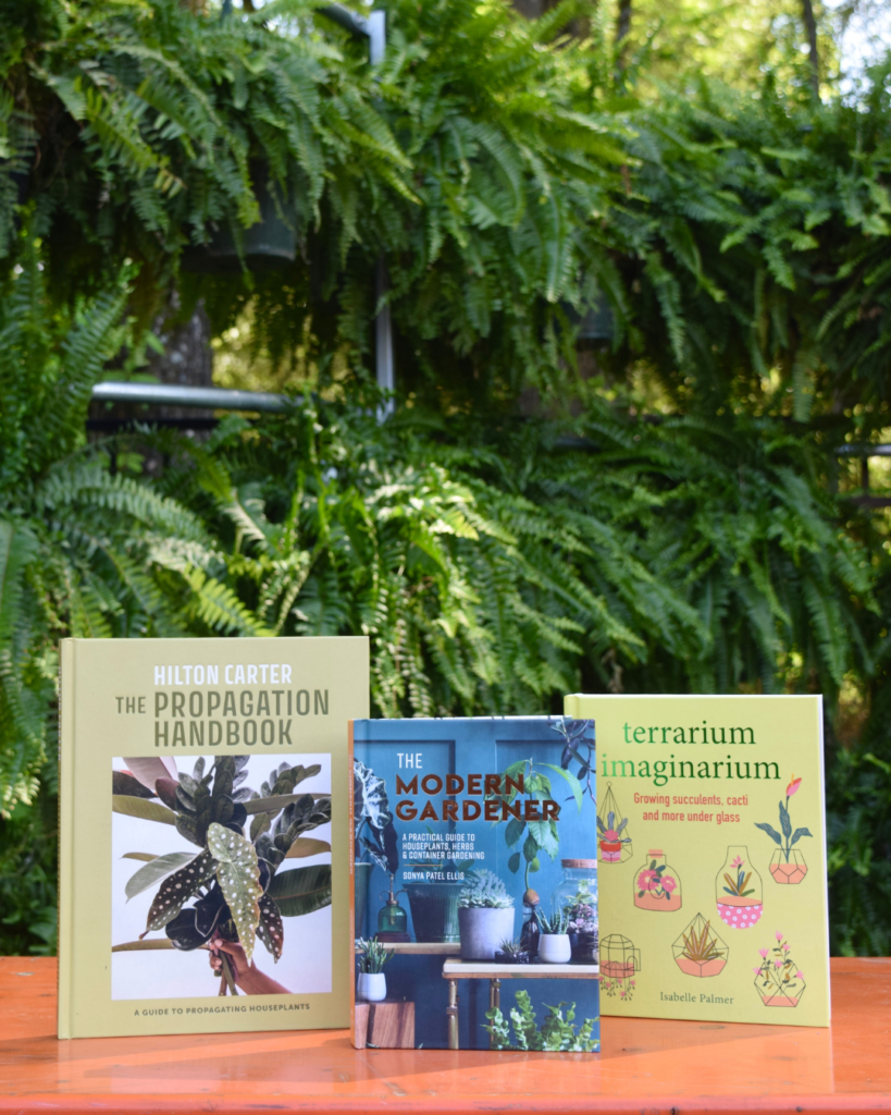Three gardening books are displayed upright on a wooden surface outdoors, with lush green ferns in the background. The book covers feature plants and illustrations related to propagation, modern gardening, and terrariums.