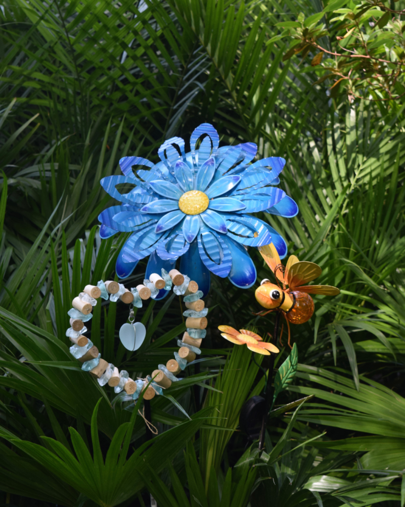 A bright blue metal flower, a heart-shaped shell decoration, and a decorative yellow bee are displayed among lush green plants and palm leaves in sunlight.