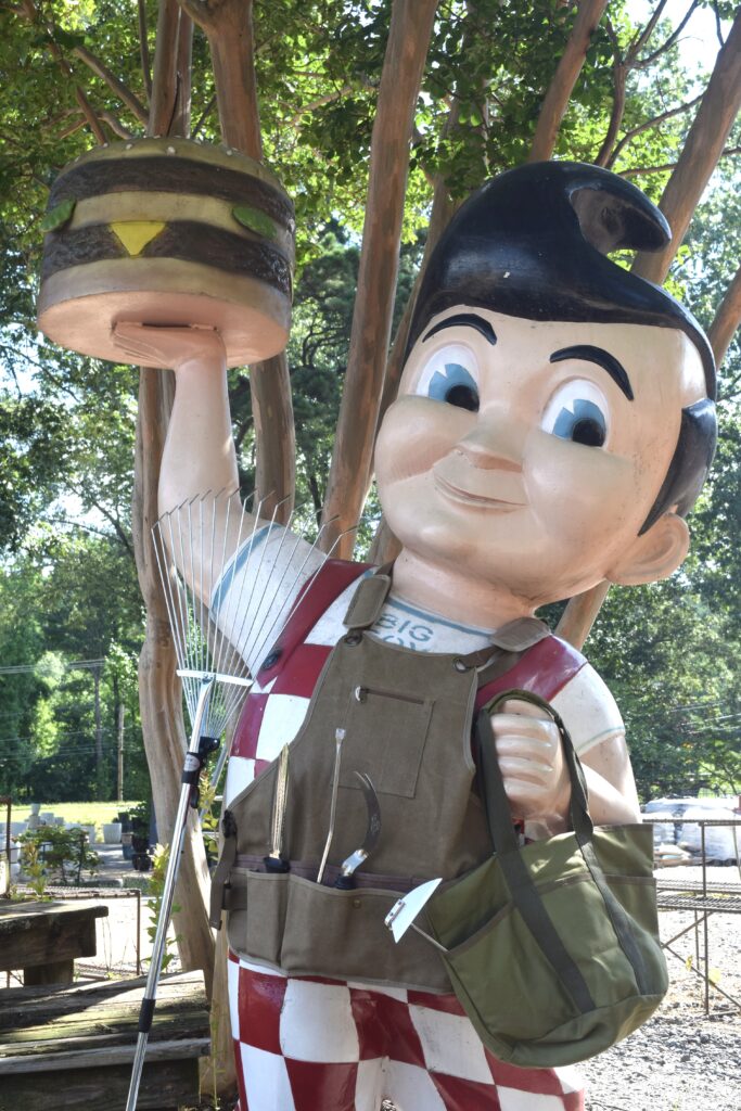 A large statue of a boy with black hair in a red-and-white checkered outfit holds a giant cheeseburger. The statue is decorated with a fishing vest, gear, and a rake, standing outdoors among trees.