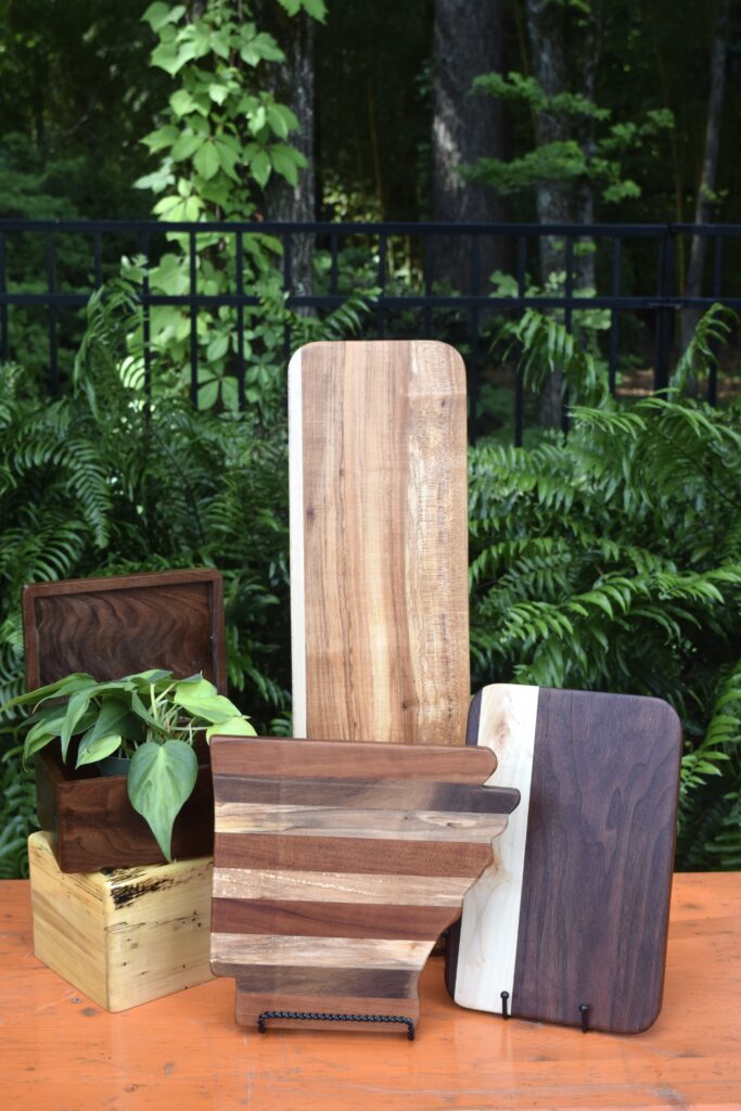 A group of wooden cutting boards and boxes of various shapes and wood tones are arranged on an orange surface, with green plants and a black metal fence in the background.