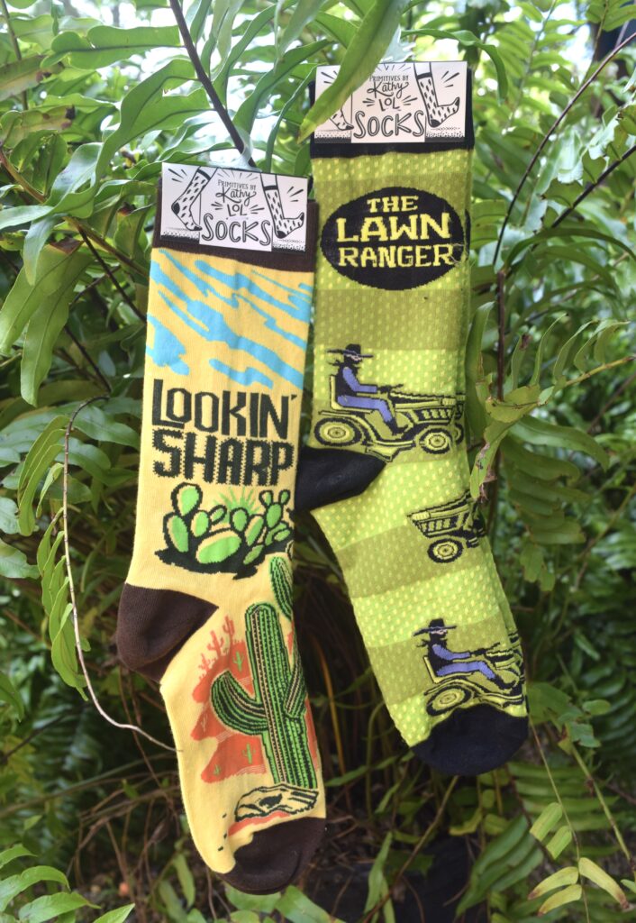 Two pairs of novelty socks hanging among green ferns. One pair is yellow with cacti and the words LOOKIN SHARP. The other is green with riding mowers and the words THE LAWN RANGER.