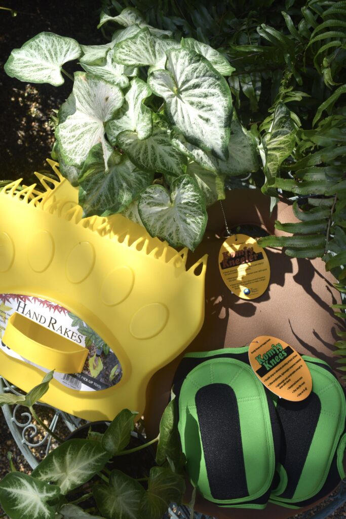 Yellow plastic hand rakes, green and black kneeling pads, and plant leaves are arranged on a round table outdoors in bright sunlight. Tags with product information are visible.