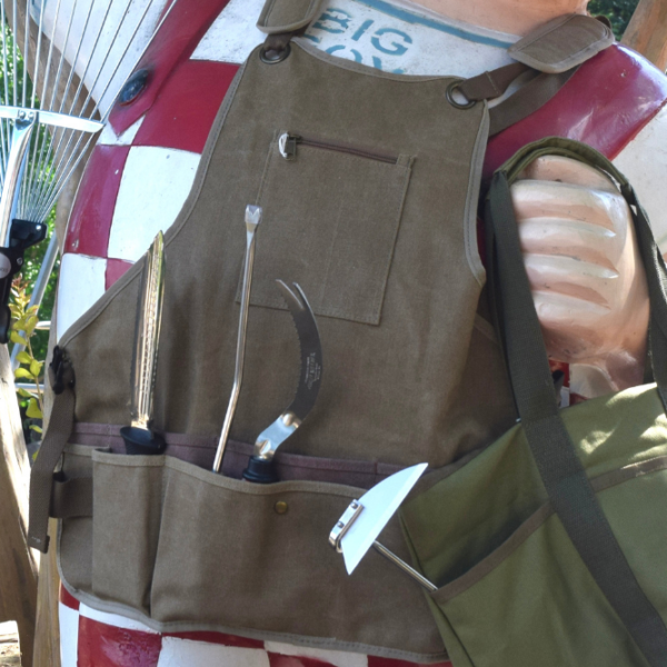 A close-up of a statue wearing a gardening apron with several garden tools in its pockets, holding a green tote bag and standing outdoors near rakes and bags of soil.